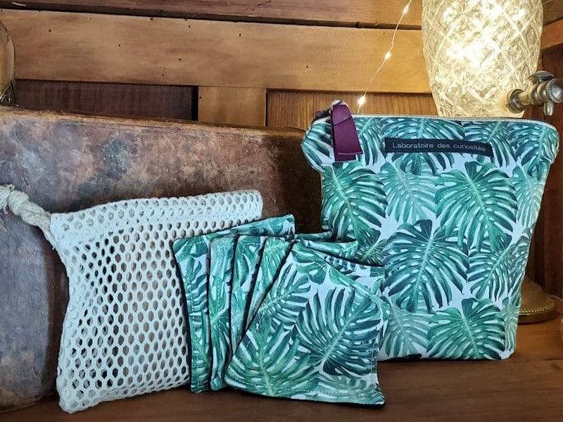 Une sélection d'alternatives durables pour la routine de beauté quotidienne. Un sac de rangement à imprimé tropical vert et blanc contient plusieurs lingettes lavables du même motif. À côté, un filet en maille de coton naturel pour y glisser son savon solide. L'ensemble repose sur un fond de bois chaleureux.